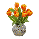 A bouquet of orange tulips with green leaves arranged in a round gray vase with gold cutout details, isolated on a white background.