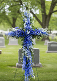 Floral cross arrangement with blue and white flowers against a beige wall.