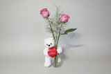 Two pink roses in a red vase with a white bear holding a red heart on a white surface.