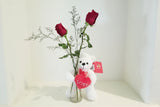 White teddy bear holding a red heart with 'LOVE' on it, standing next to a vase with red roses on a white background.