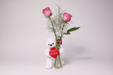 White teddy bear holding a heart with 'LOVE' text, standing next to a red vase with pink roses on a white surface.