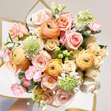 A bouquet of flowers featuring roses, ranunculus, and other seasonal flowers in pink, peach, and cream colors, with greenery.