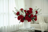 Bouquet of red roses and white roses in a white vase on a table with a white curtain and sofa in the background.