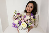A floral arrangement with a variety of flowers in shades of purple, pink, white, and green, presented in a white purse-like container.