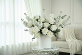 White floral arrangement in a vase on a small table with a white curtain and sofa in the background