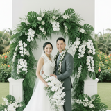 A bride and groom standing together in front of a lush tropical floral arch. The bride wears a sleeveless white wedding gown and white flower lei, holding a large white orchid bouquet.