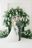 A smiling bride in a white lace wedding gown with a floral lei stands beside a groom in a gray suit and light blue tie, both wearing green and white flower lei, in front of a white floral backdrop.