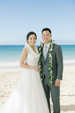 A smiling bride in a white lace wedding gown and lei stands beside a groom in a gray suit and floral lei on a sunny beach with turquoise ocean water and pale sand in the background.