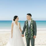 A bride in a white lace wedding gown and a groom in a gray suit holding hands on a sandy beach, with the ocean and blue sky behind them. The groom wears a green and white floral lei, and the bride wears a matching lei.
